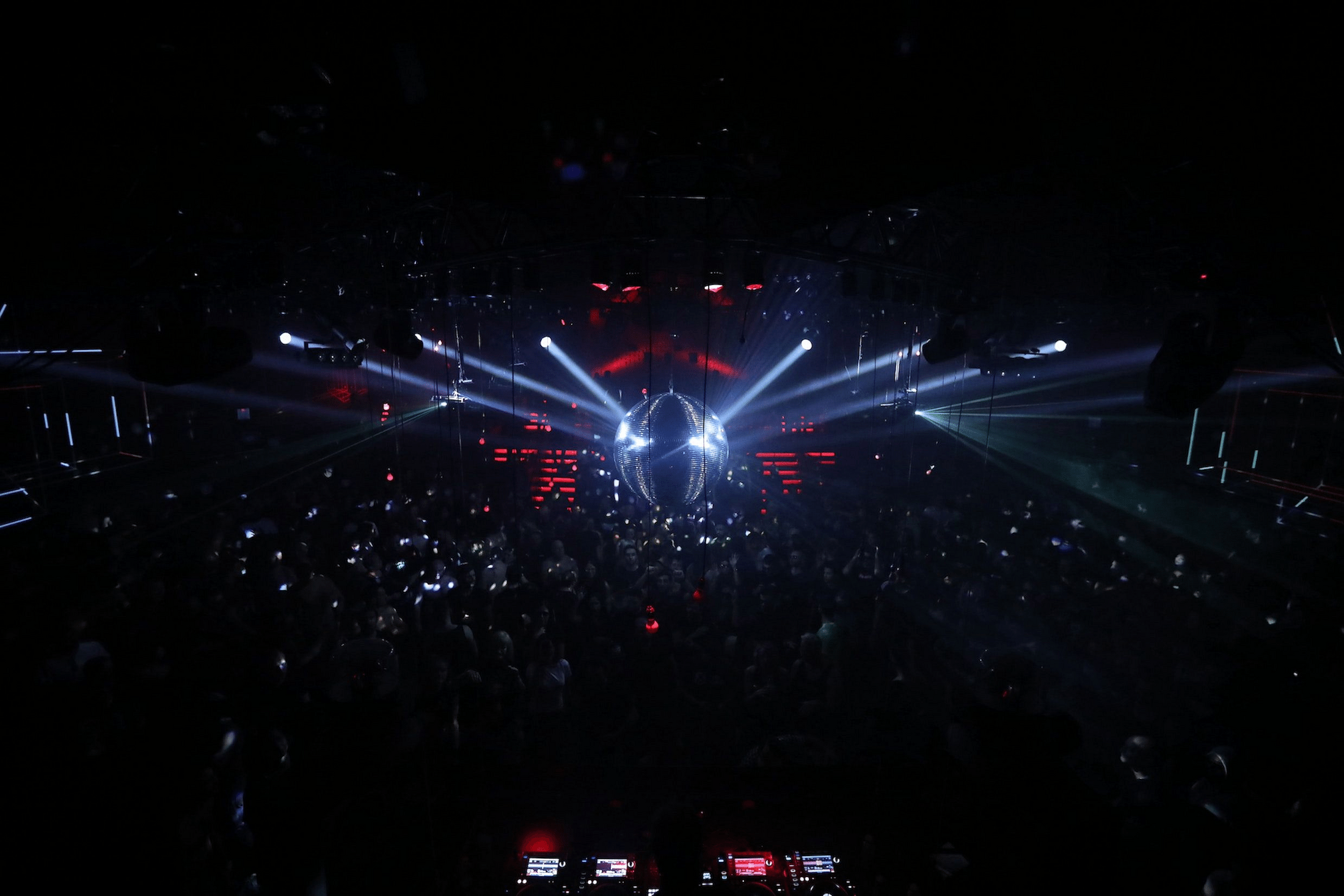 A dark, packed dance floor lit by white spotlights off a disco ball, red panels glowing in the distance